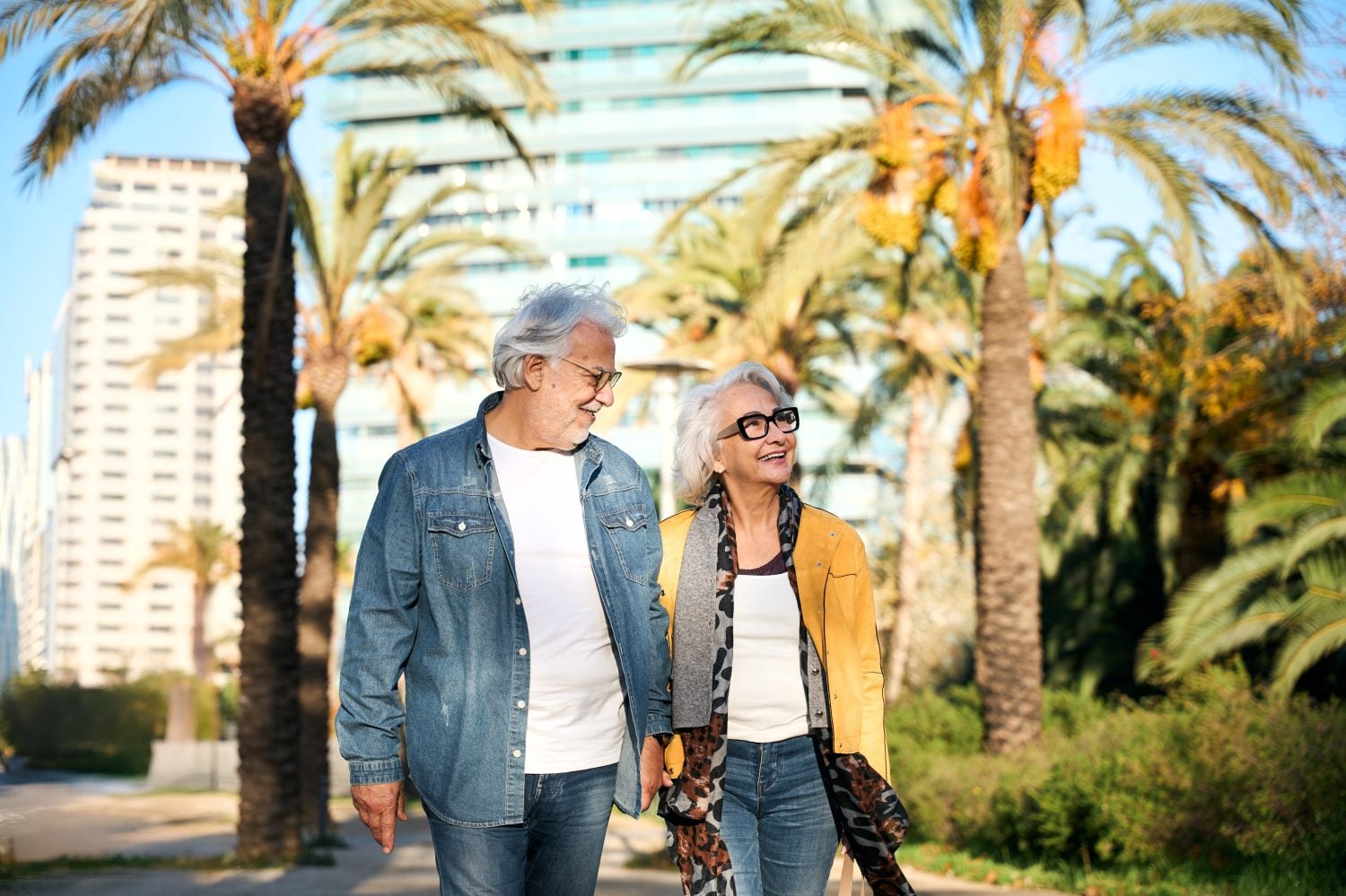 An older man and woman, both with gray hair and wearing glasses, walk side-by-side on a paved path, smiling and looking upwards. The man wears a denim shirt over a white t-shirt and jeans. The woman wears a yellow jacket, a white top, jeans, and a patterned scarf. In the background are tall palm trees and modern buildings under a bright blue sky.
