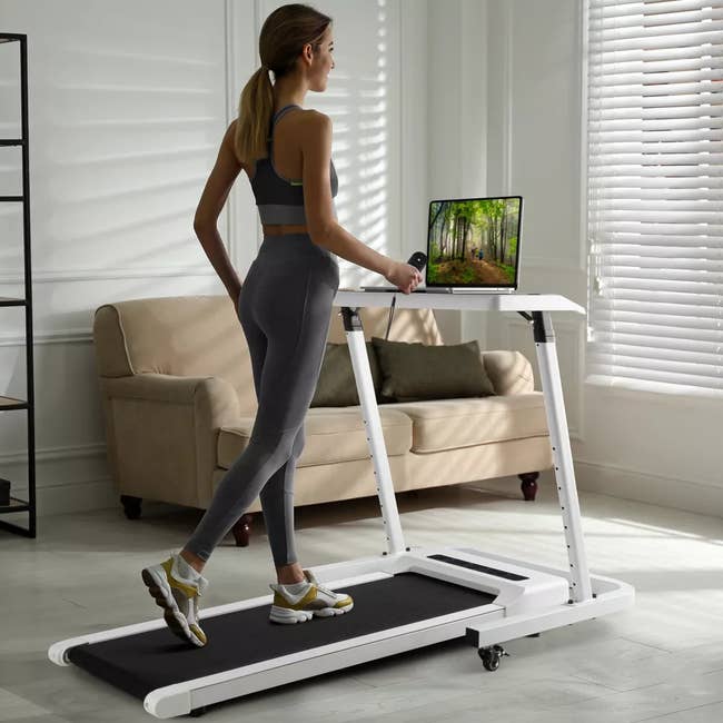 Person walks on a treadmill desk while holding a mug, with a natural scene displayed on its screen