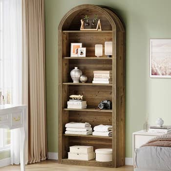 Wooden arched bookshelf in a bedroom, styled with decor, books, vases, and neatly folded towels, next to a window with curtains