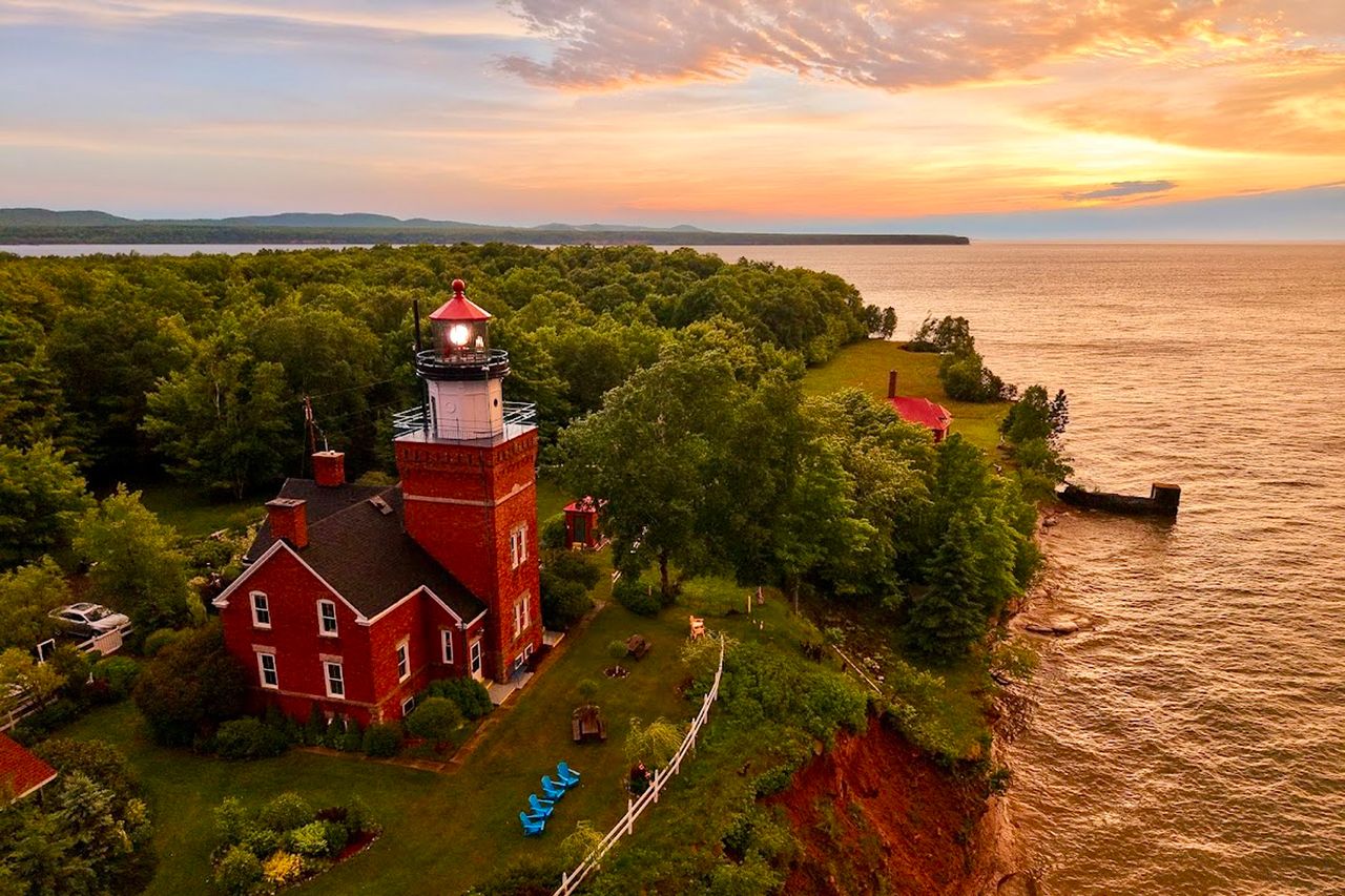 A lighthouse on a cliff overlooking a body of water with a scenic background of trees and a sunset horizon