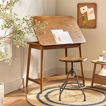 Adjustable wooden drafting table with a stool and drawing tools, situated in a cozy room with a small plant and a cork board on the wall