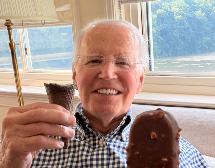 An elderly man with glasses smiles while holding an ice cream cone and bar in a room with a window view of greenery outside