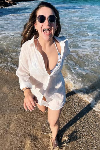 A reviewer in sunglasses and a white beach cover-up walks along the shoreline, smiling with their tongue out. The ocean waves are in the background