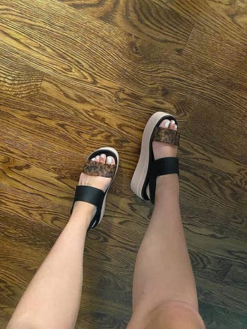 Sandals with leopard print straps and platform soles worn on feet standing on a wood floor