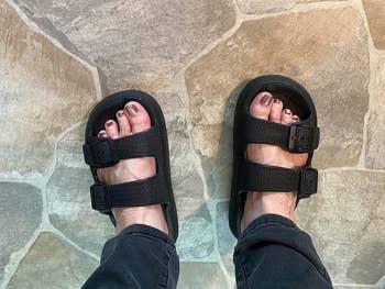 Feet in black sandals with two straps and buckles, worn with casual pants, standing on a stone tile floor