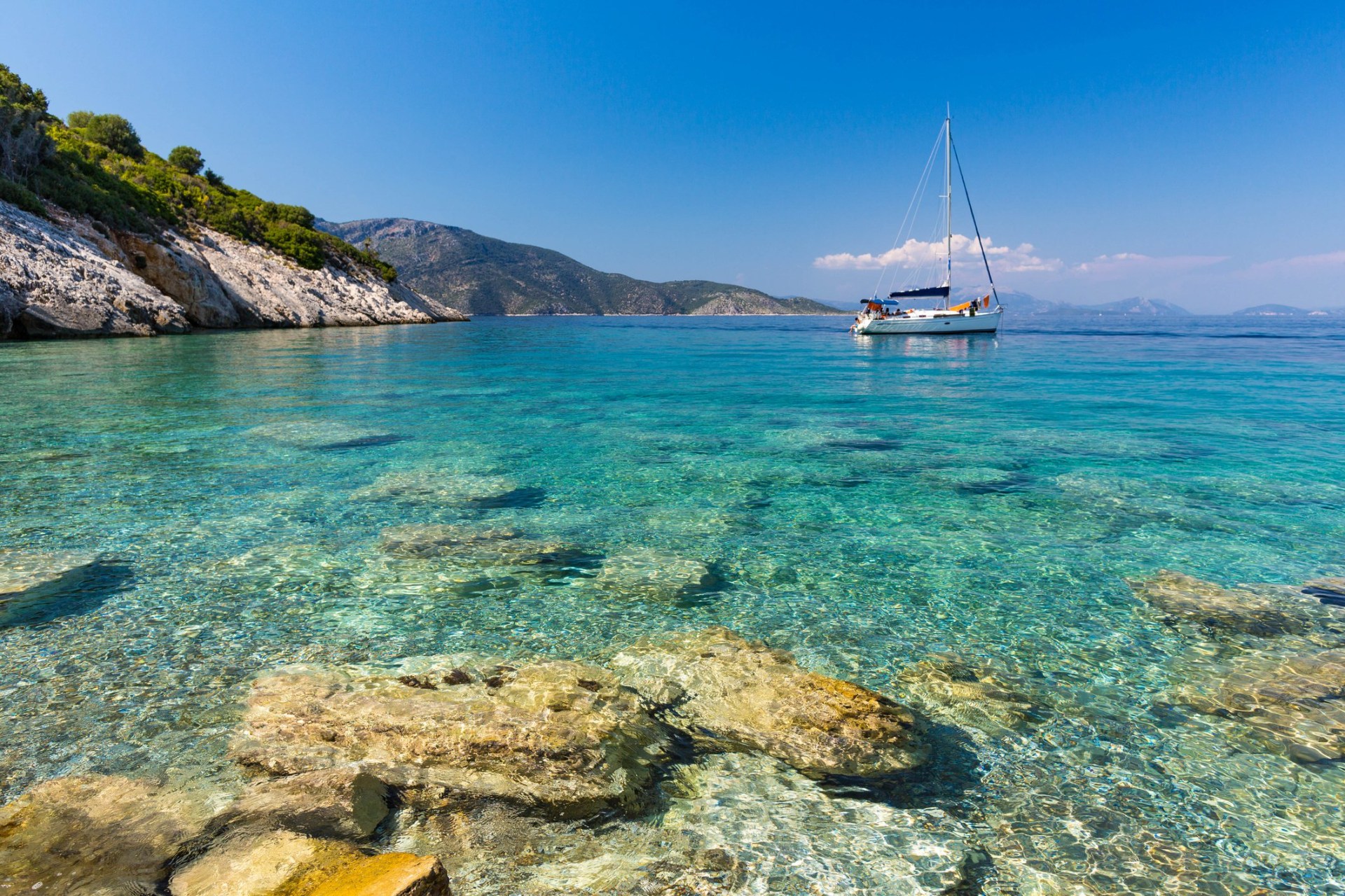 A sailing boat seen from the turquoise sea in Ithaca, Greece, with grassy cliffs behind it.