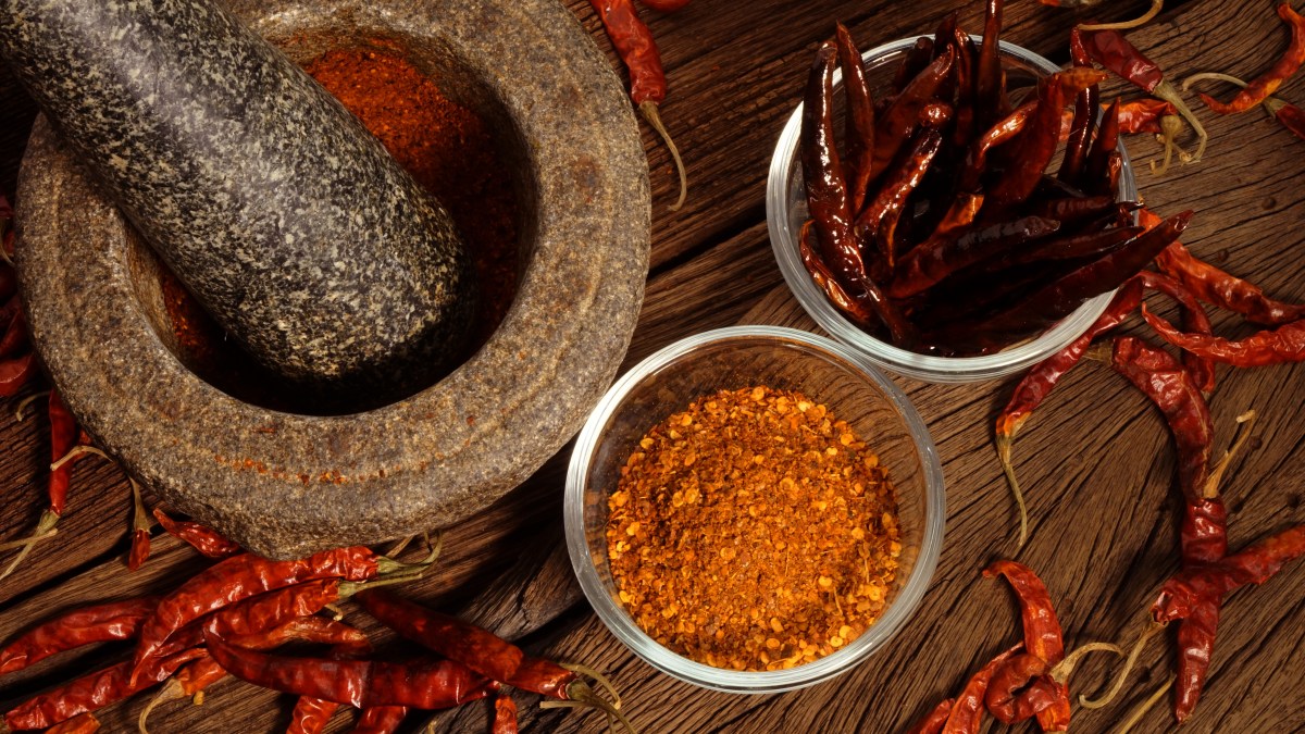 Dried red chilies sit next to a stone mortar and pestle, next to a small bowl of ground chili powder.