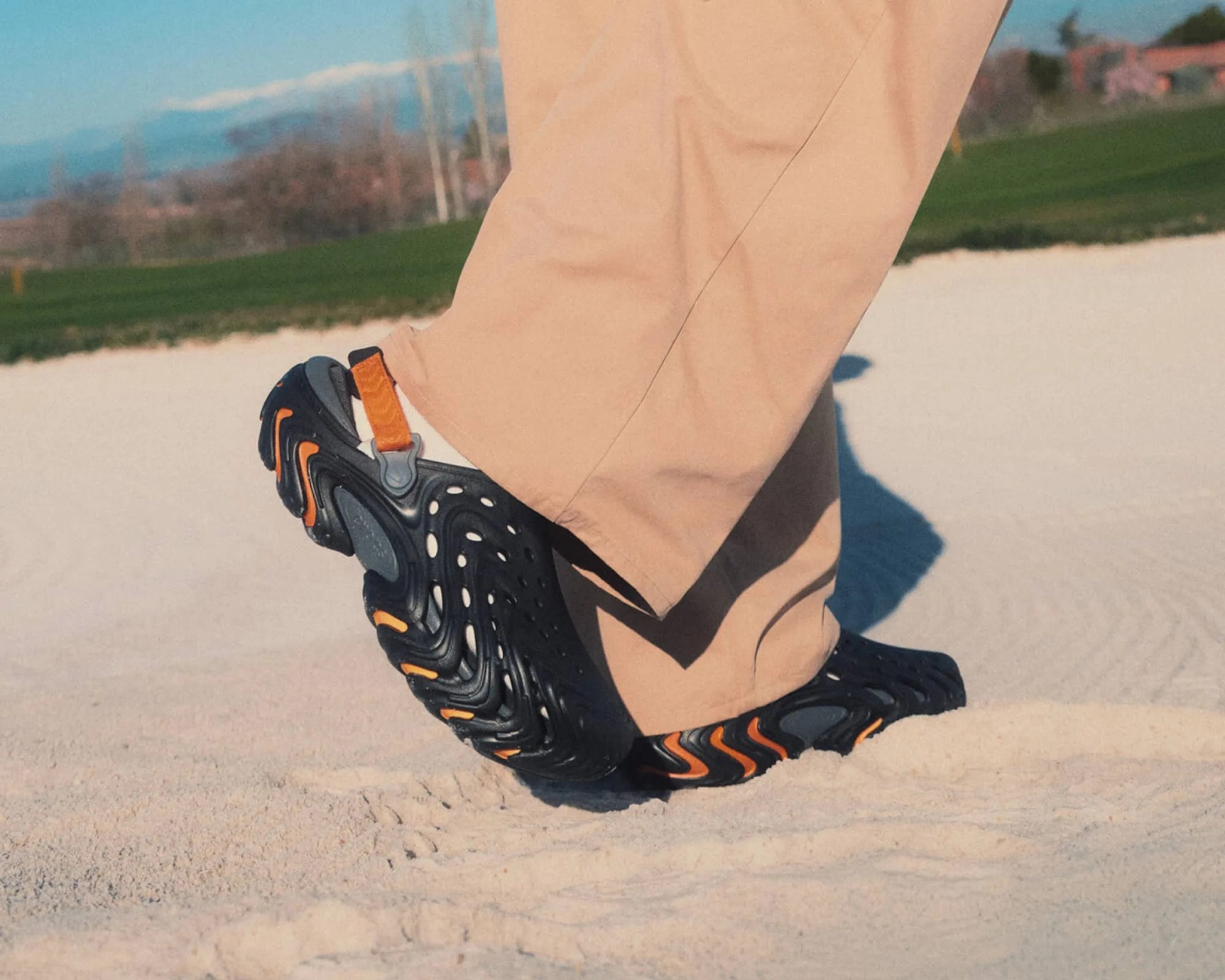 Black and orange athletic shoes with a unique wavy sole worn with beige pants on sandy ground.