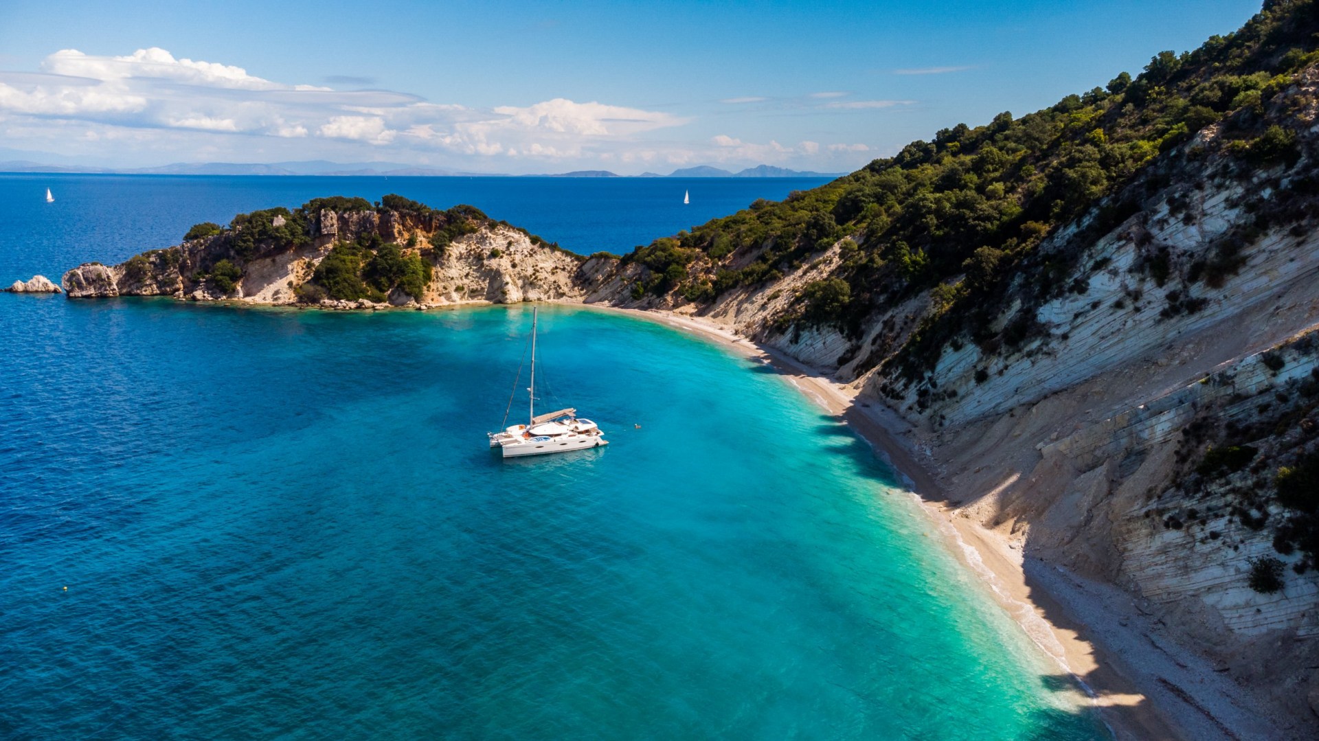 A lone white boat sitting on the calm turquoise water of Ithaca, Greece, surrounded by a cliff and a sandy cove. The sky above is light blue with a few small clouds.