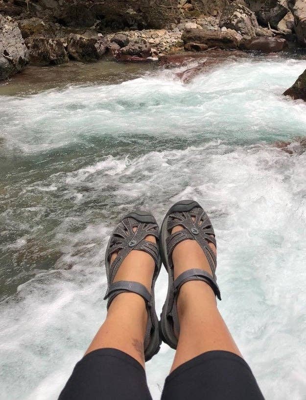 Person sitting by a rushing river with feet wearing stylish open-toe sandals, suggesting comfort and adventure in outdoor settings