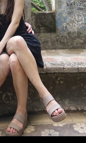 A person sits on a bench wearing platform sandals and a casual dress, with their legs crossed. The setting appears relaxed and informal