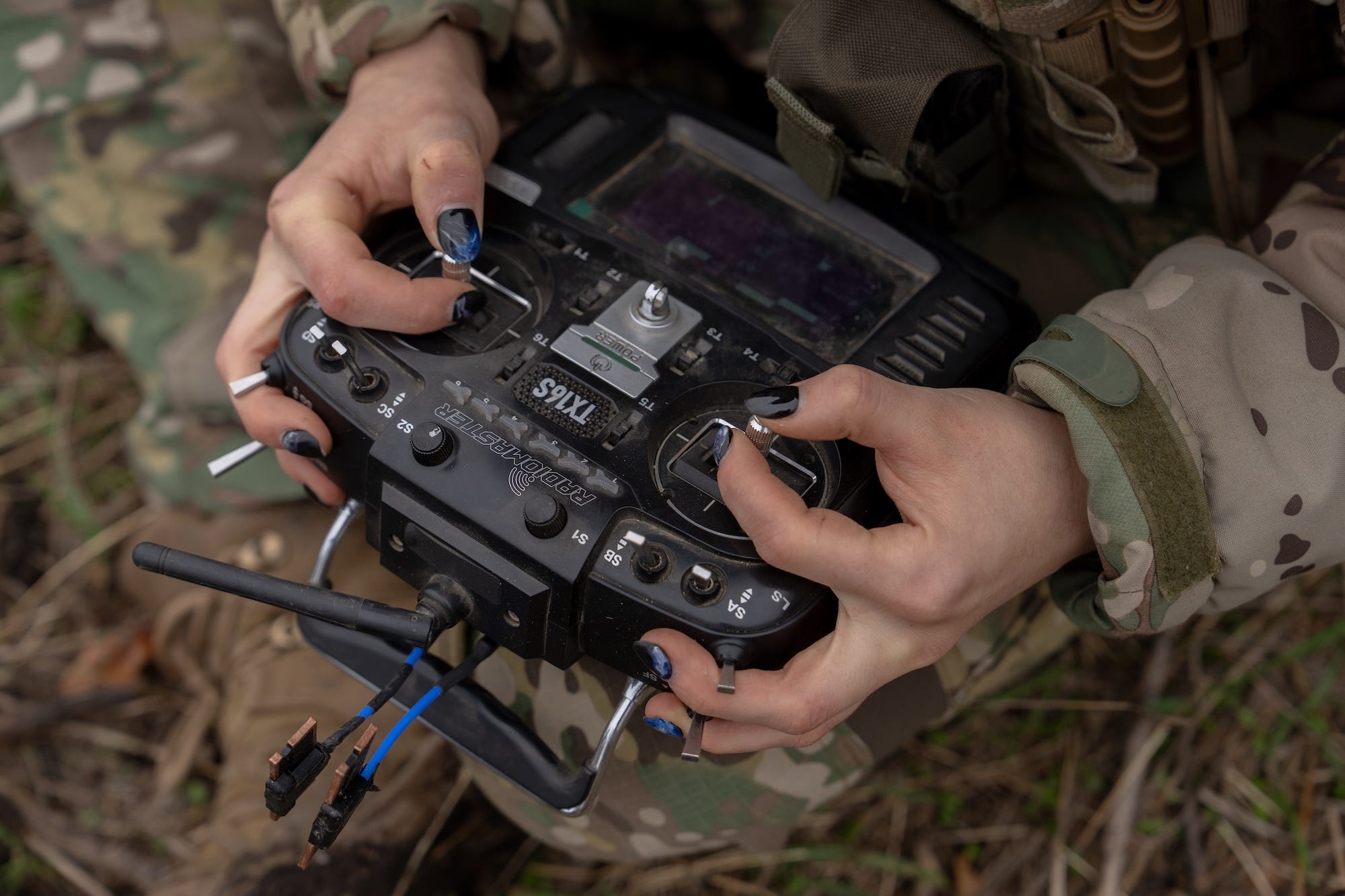 Hands in camouflage clothing operate a RadioMaster TX16S remote controller outdoors among grass and leaves.
