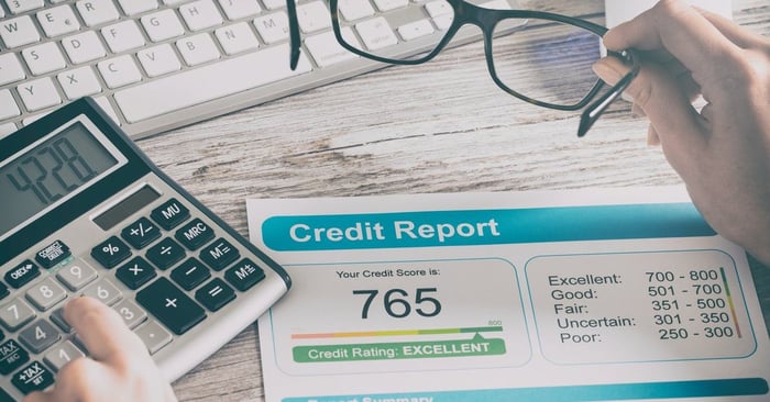 Credit report that reads 765 on a desk where someone is typing on a calculator and their holding glasses