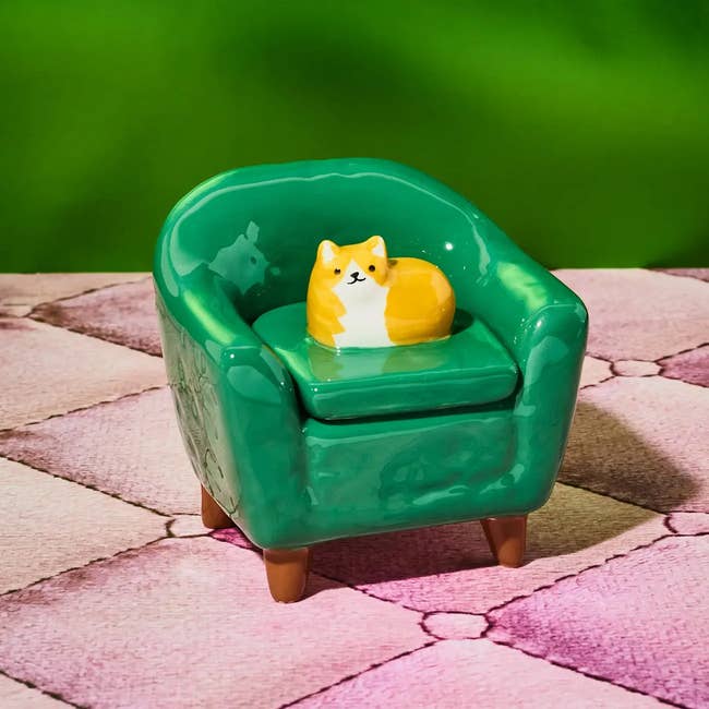 Ceramic figurine of a cat resting on a green armchair, placed on a patterned surface. Perfect for home decor or as a gift for cat lovers
