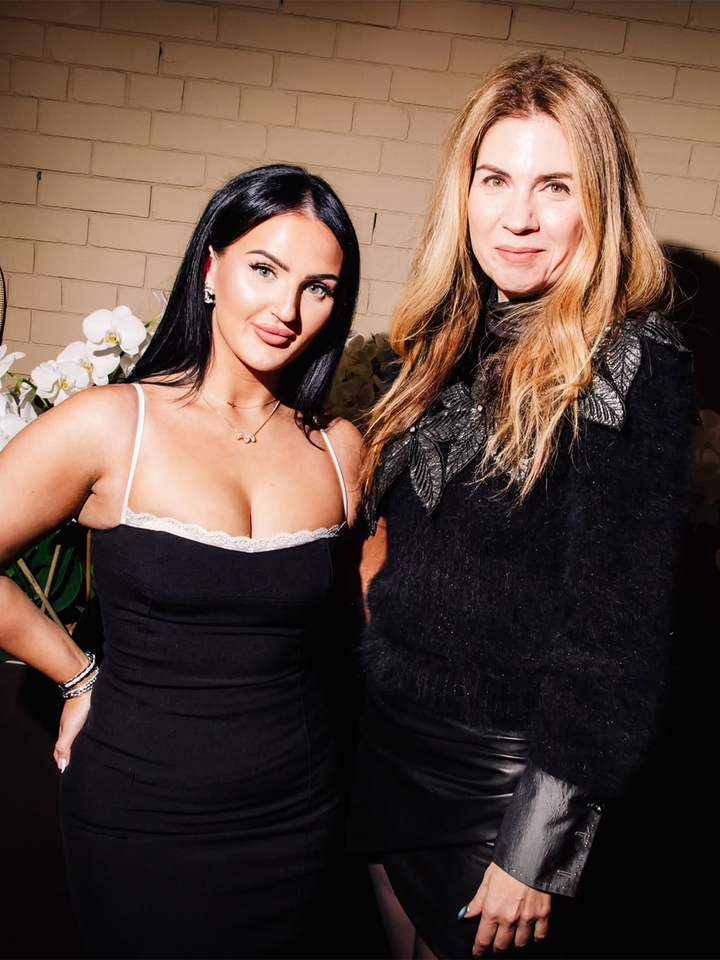 Two women posing together indoors one with dark hair and the other with light hair in front of a wall and flowers