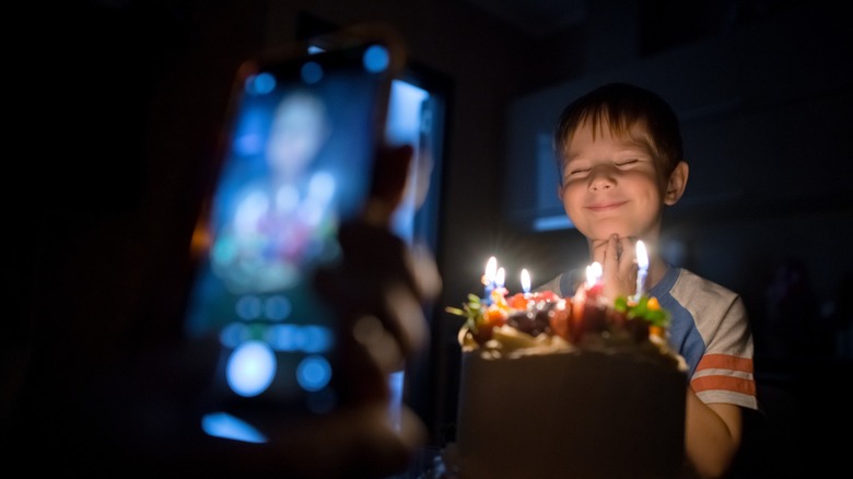 A phone takes a picture of a young child illuminated by candles on a cake.