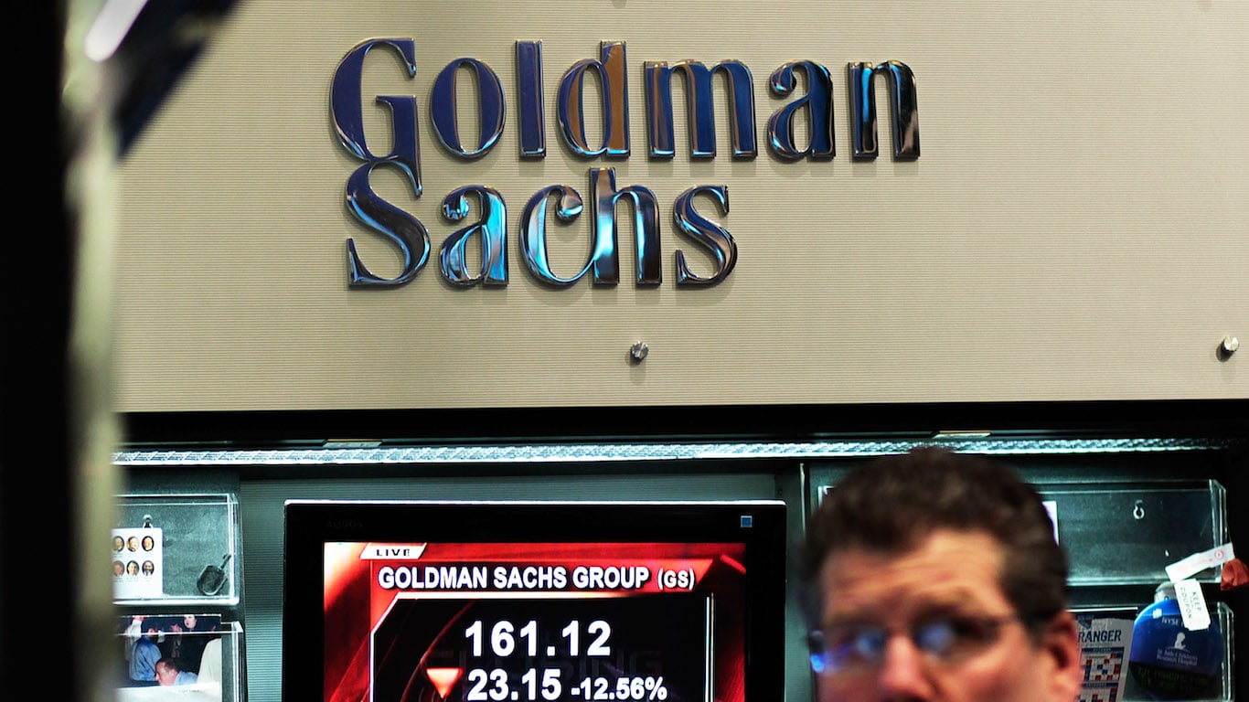 The polished, metallic 'Goldman Sachs' logo is mounted on a light grey, horizontally-textured wall. Below the logo, a digital screen displays 'GOLDMAN SACHS GROUP (GS)' with a stock price of '161.12' and a significant decrease of '23.15 -12.56%'. The blurred upper part of a person's head and shoulders, wearing glasses, is visible in the lower right foreground.