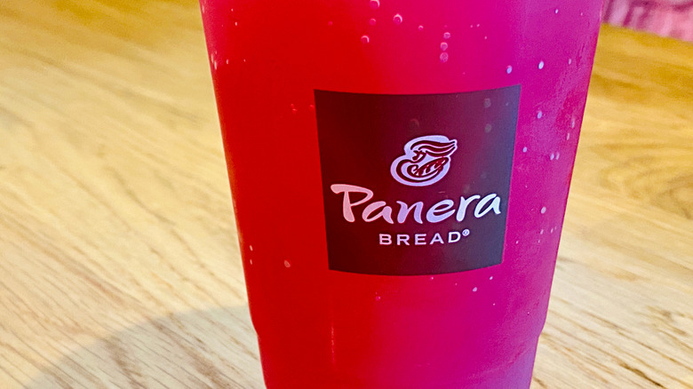 A Panera Bread branded plastic cup filled with a red-pink beverage sits on a wooden table