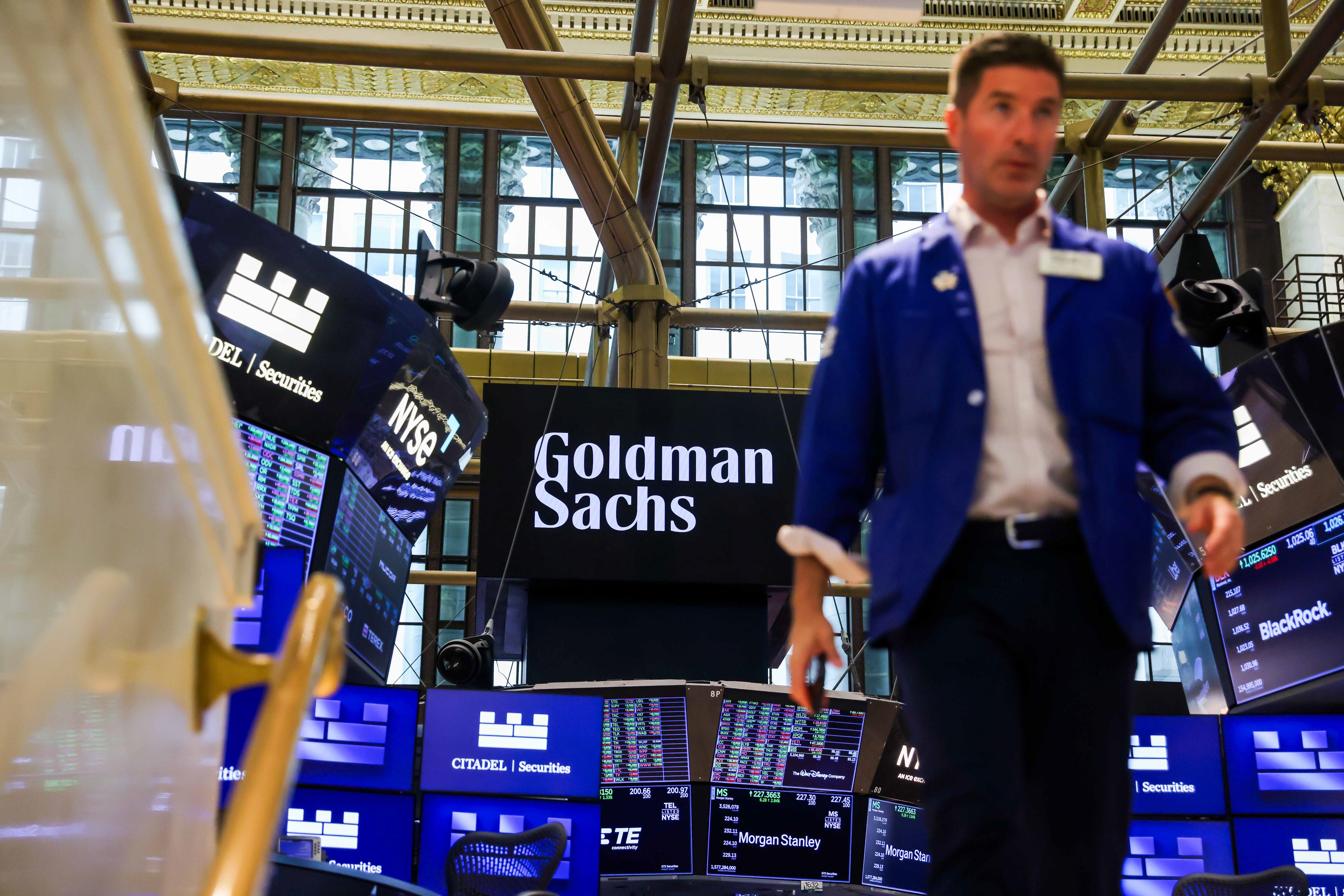 Blue-jacketed floor worker passes trading screens showing Goldman Sachs and other financial firm names on an exchange floor.