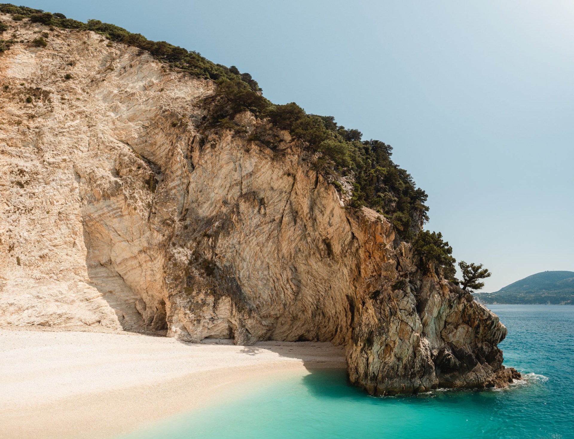 A quiet white sand beach in Ithaca, Greece. There is a large cliff by the sand, and bright turquoise water. The sky is light blue.