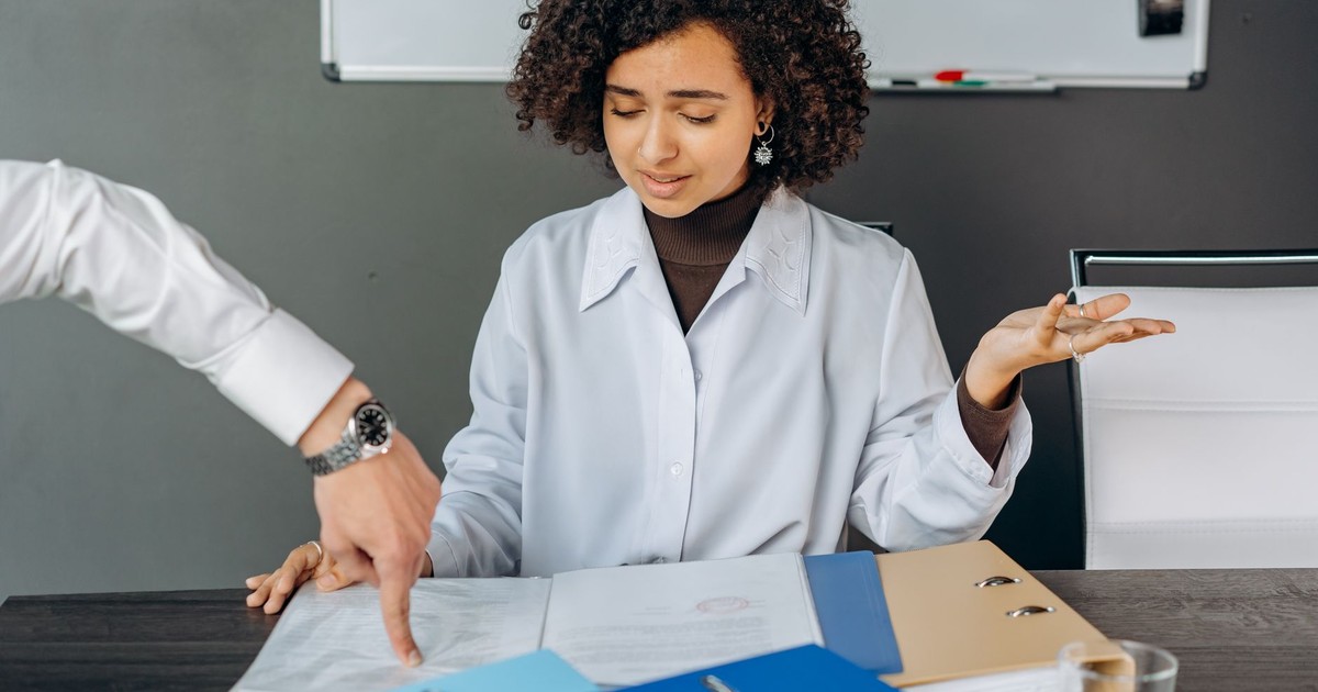 A curly-haired woman is overwhelmed by paperwork in an office setting, multitasking with documents and files.