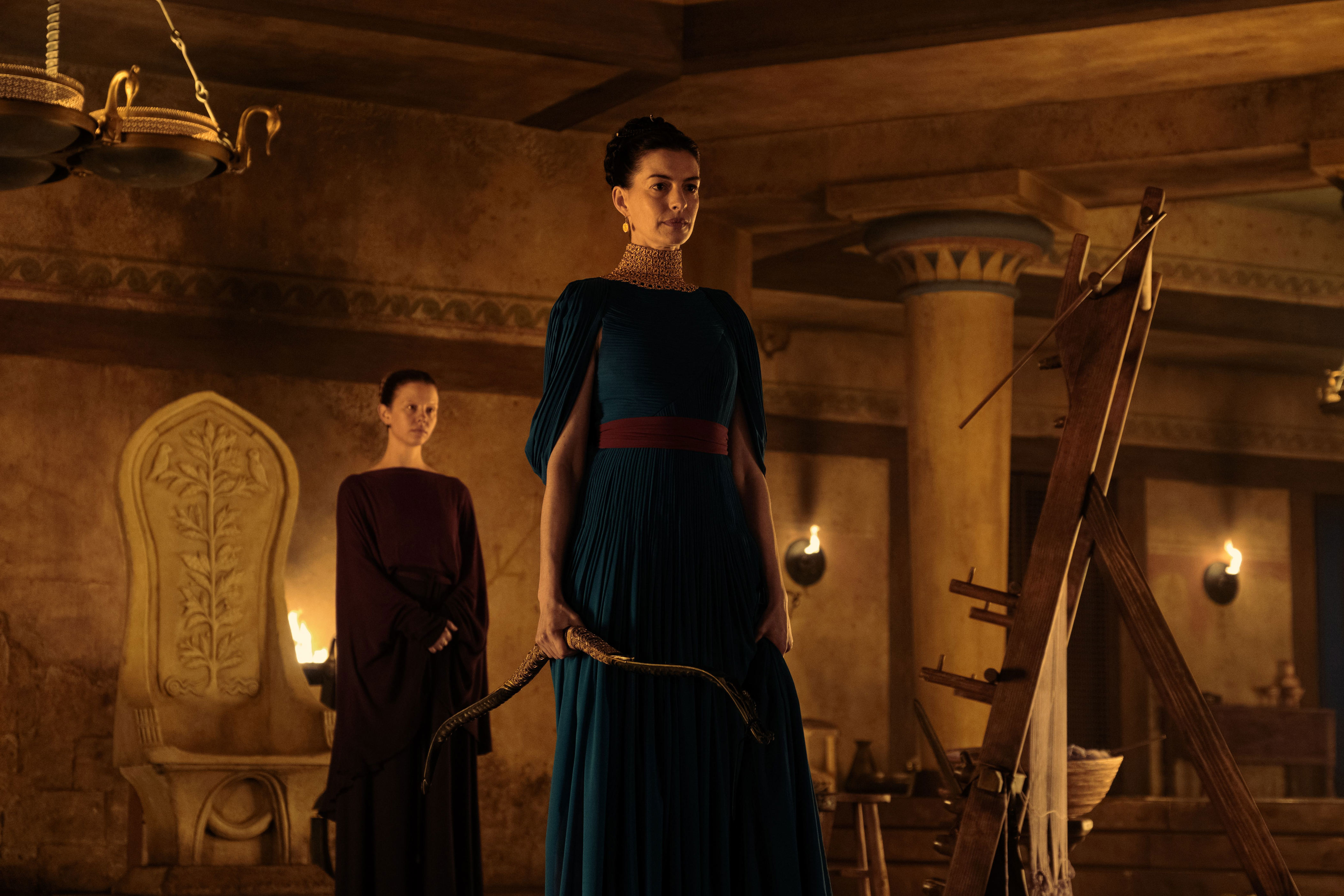 Anne Hathaway, as Penelope, stands in front of her servant Melantho, played by Mia Goth.