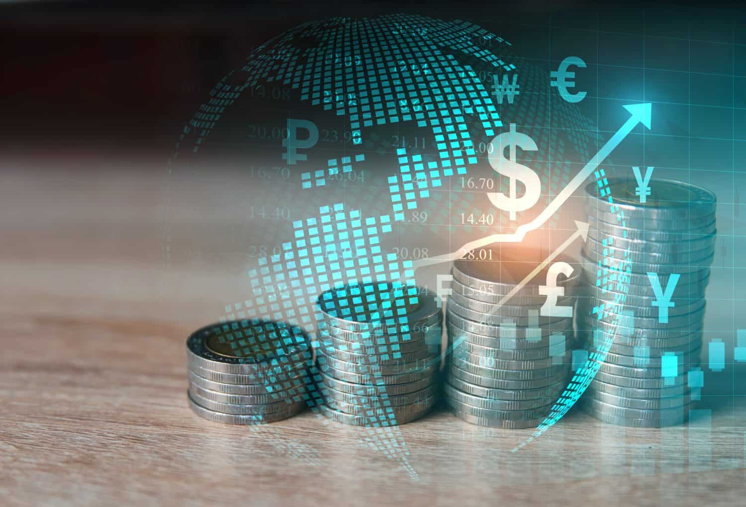 Four stacks of silver coins of increasing height are arranged on a light wooden surface, set against a dark, blurred background. A semi-transparent blue digital grid pattern of a world map overlays the scene, along with various white currency symbols including Dollar, Euro, Pound, Yen, and Ruble. A prominent white arrow graphic curves upwards from left to right, indicating growth, with numerical data points scattered across the digital map, and a bright light emanating near the peak of the arrow.