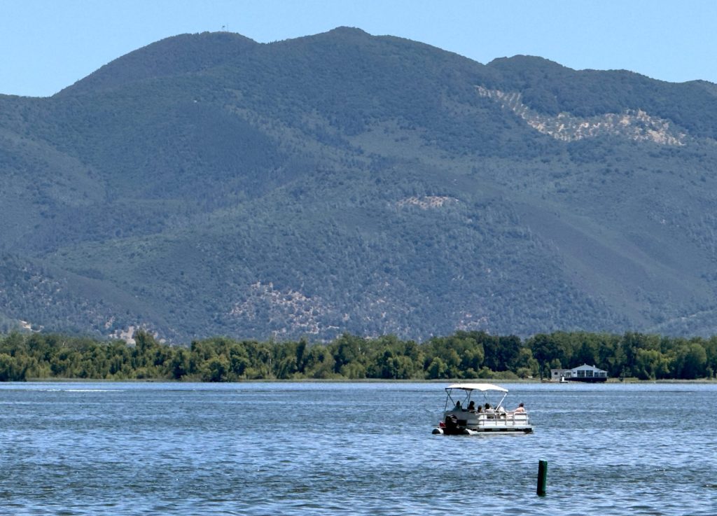 A pontoon boat with passengers glides across a calm lake, with lush green mountains rising in the background under a clear blue sky.