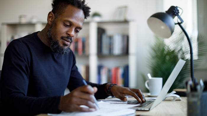 A young investor is sitting at a desk while reviewing printed documents and using a laptop.