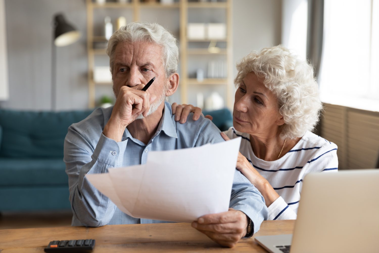 A concerned senior man with gray hair and a beard, wearing a light blue checkered shirt, holds papers and pensively touches his chin. Beside him, an older woman with curly white hair, wearing a striped shirt, places a comforting hand on his shoulder and looks at him with a worried expression. They are seated at a wooden table with a calculator and a laptop, suggesting they are reviewing financial documents in a home setting. The mood is serious and contemplative.
