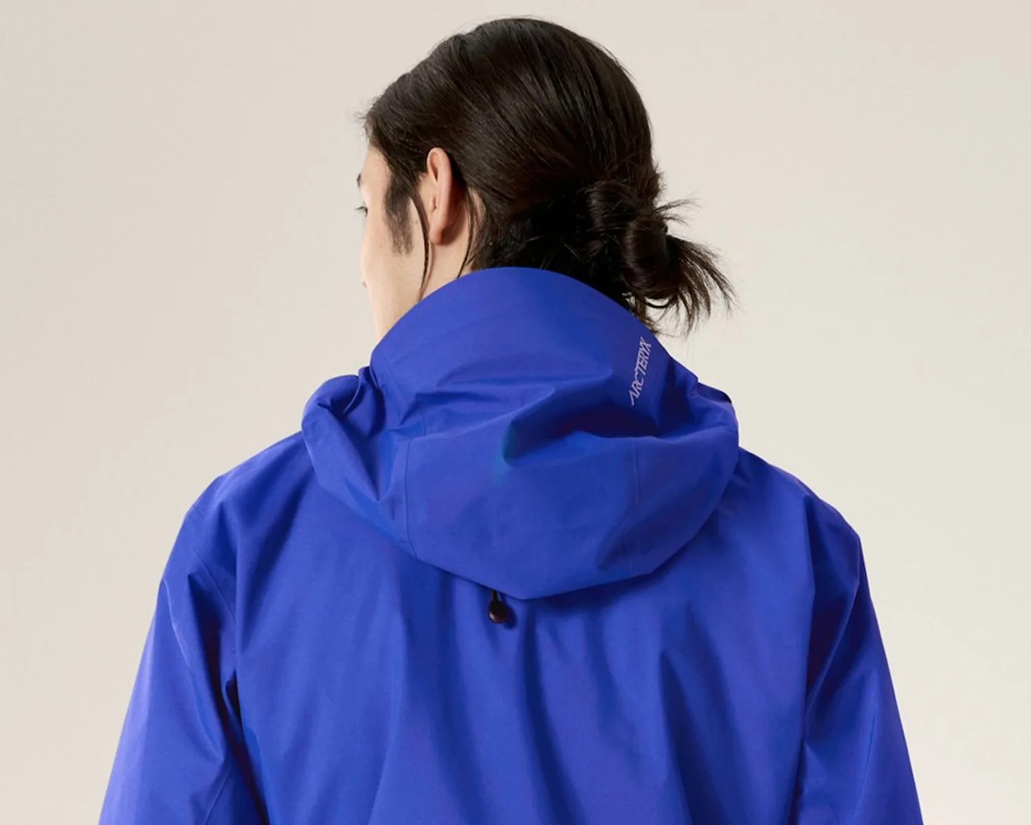 Bright blue Arc'teryx jacket with hood worn by a person with dark hair tied in a low bun, shown from the back.