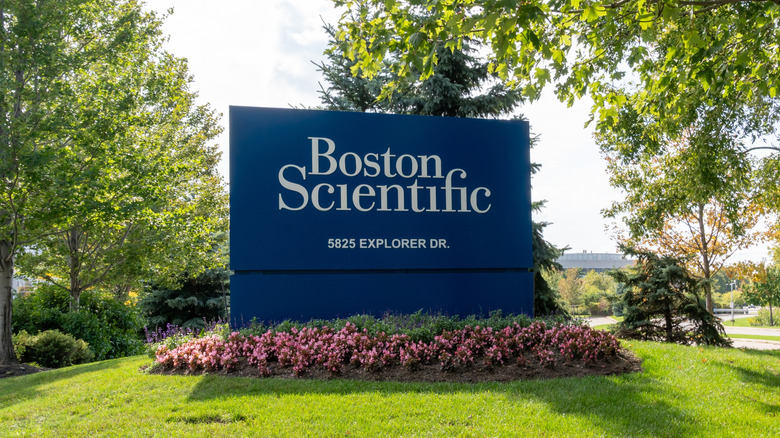 An outdoor Boston Scientific sign with an address listed on it surrounded by pink flowers at its base