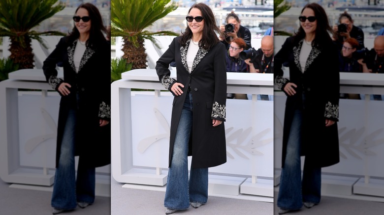 Julia Louis-Dreyfus posing at an event with her hand on her hip wearing flared jeans, a black coat, and dark sunglasses.