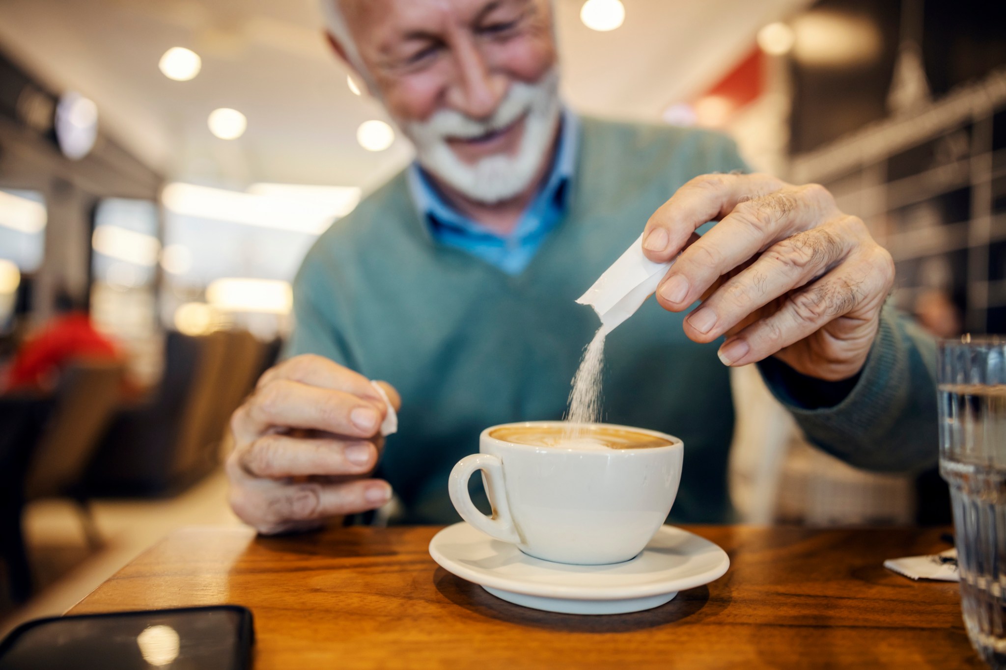 Selective focus on old man’s hands adding bag of sugar into coffee at cafeteria.