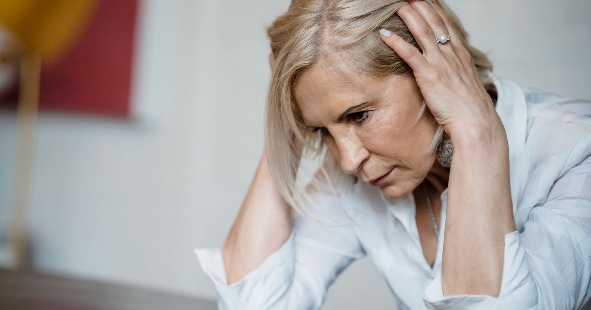 Woman with long blonde hair, deep in thought, wearing a white blouse indoors.