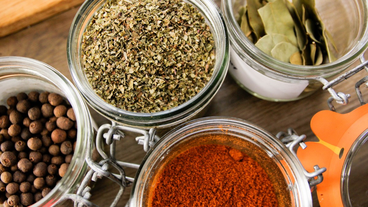 Bay leaves, oregano, allspice and paprika in glass jars with their lids open.