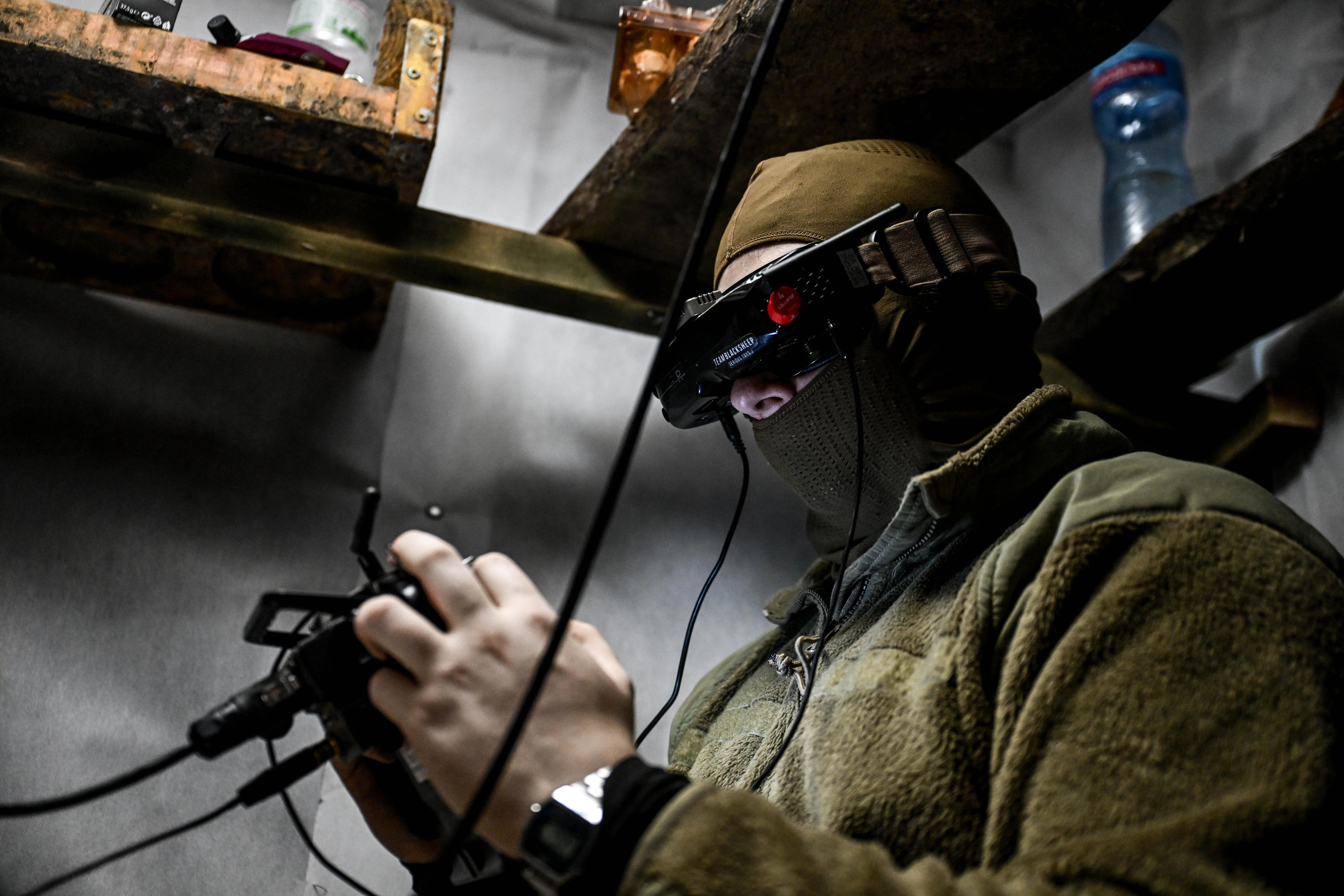 Masked person in tactical-style clothing operates a wired handheld controller while wearing electronic goggles indoors.