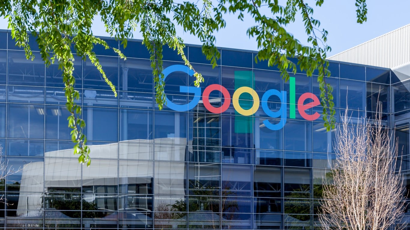 The colorful Google logo, spelling 'Google' in blue, red, yellow, and green, is displayed prominently on the reflective glass facade of a large, modern corporate building. Lush green leaves from an overhead branch partially obscure the top left corner, while reflections of other structures and the sky are visible in the glass panels. A bare-branched tree stands in the lower right foreground.
