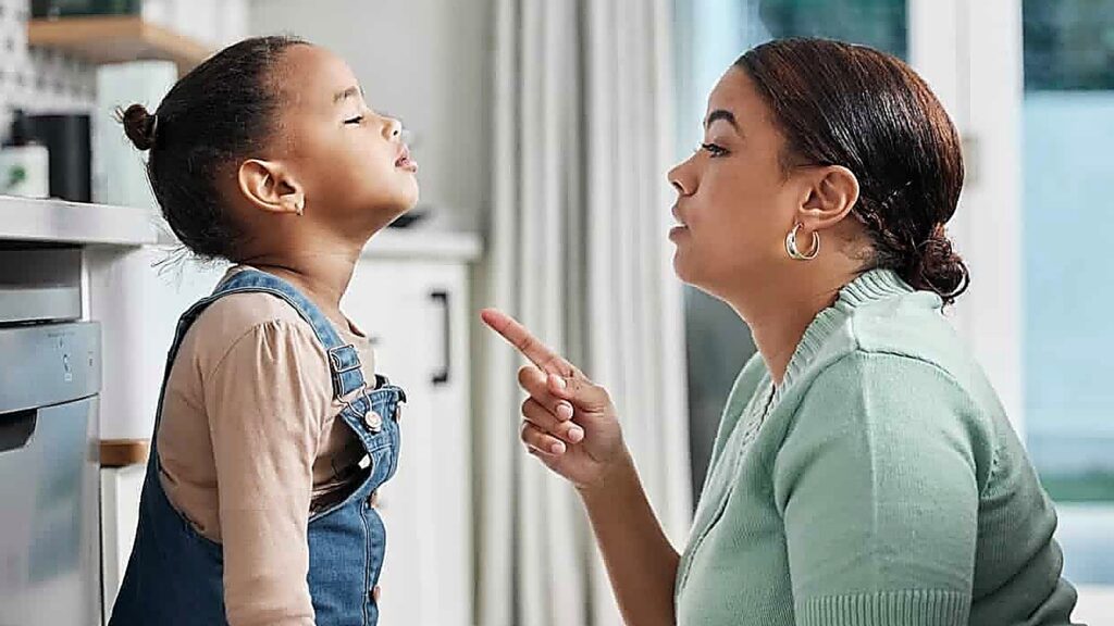 An adult is pointing a finger and talking to a young girl, who is looking up with her chin raised in a kitchen setting, capturing the tension of a power struggle between them.