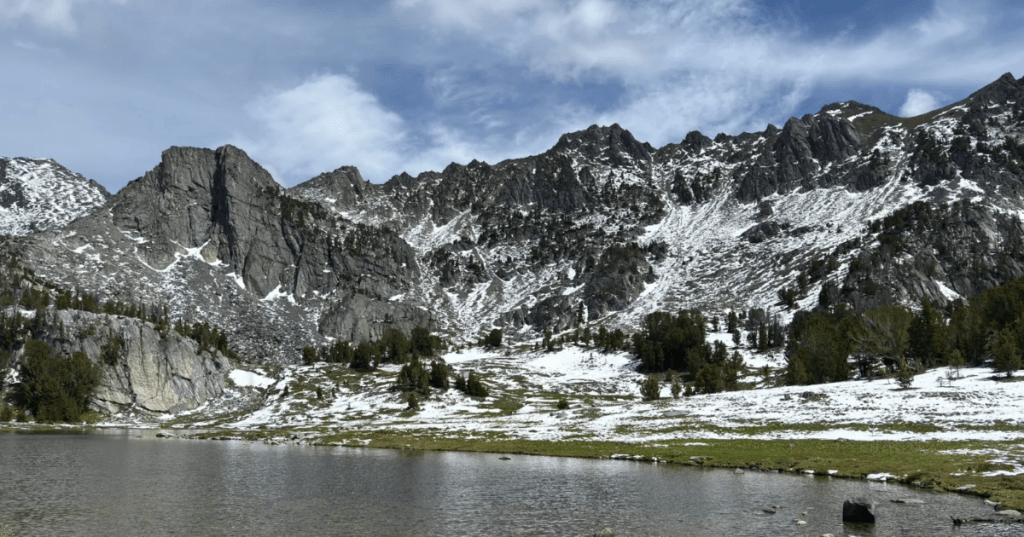 Mountains with scattered snowfall tower over a quiet lake along the Beehive Basin Trail in Montana.