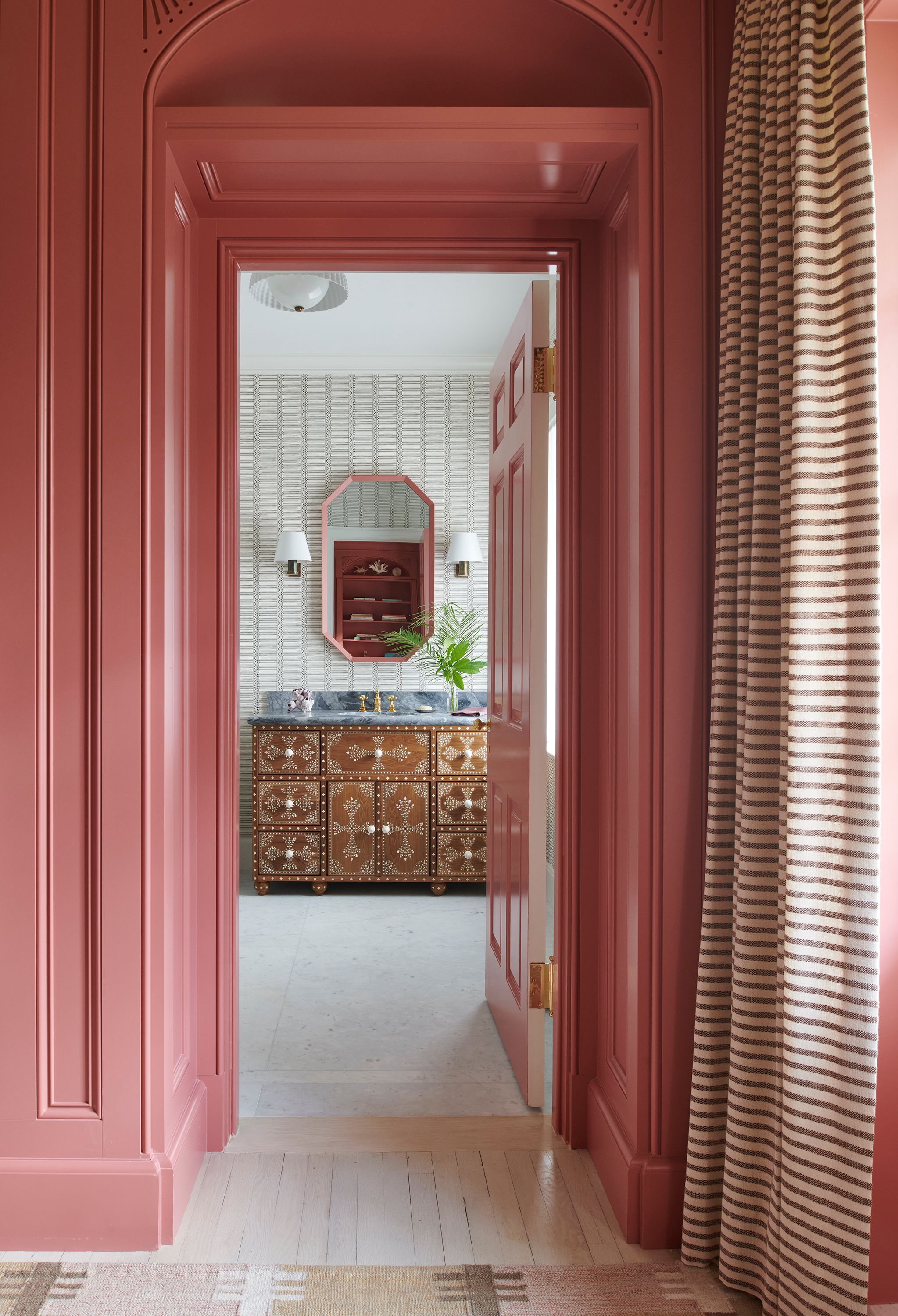 The raspberry-hued “pajama lounge” leads to another powder room hung with a matching mirror from RT Facts above a studded chest repurposed as a washstand. The light is from Urban Electric and the grass-cloth wallpaper is from Pierre Frey. The rug in the foreground is from Rug and Kilim.