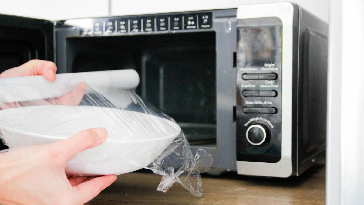 A plastic-wrapped white bowl going into a black and silver microwave.