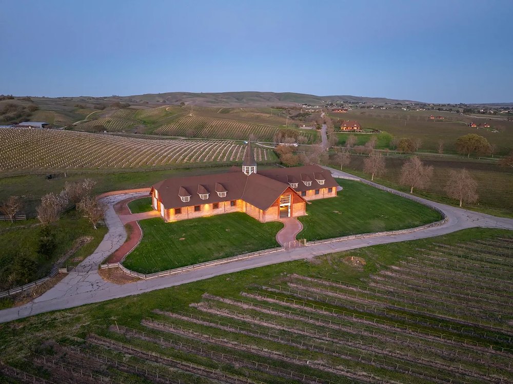 Aerial view at twilight of a grand estate and stable surrounded by vineyard rows and rolling hills.