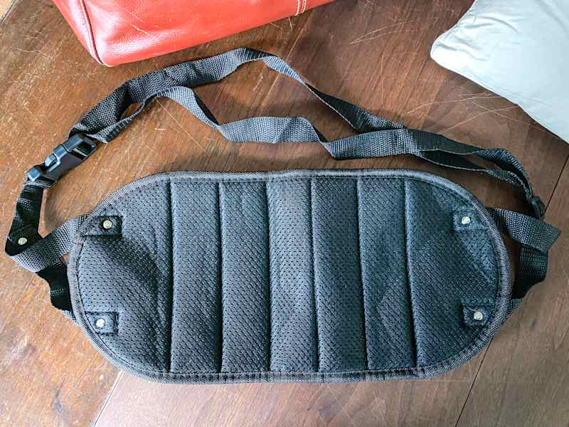 Black padded shoulder strap with adjustable clips, resting on a wooden surface near a red bag and a gray pillow.