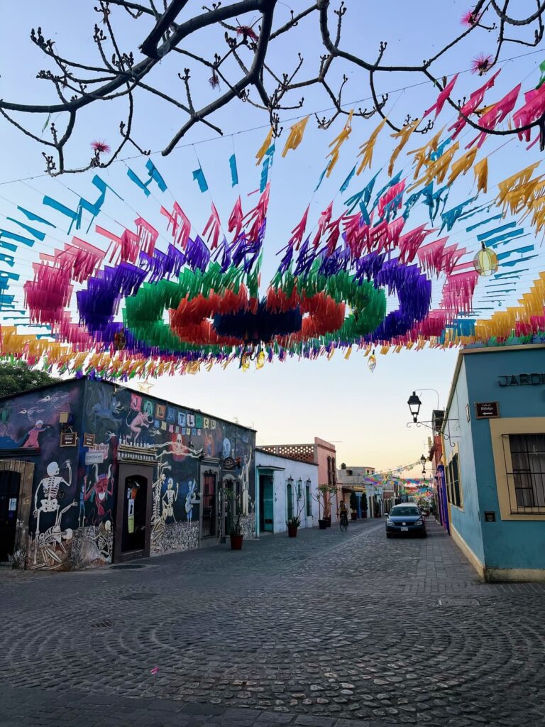 Rows of colorful papel picado banners stretch across a quiet cobblestone street lined with brightly painted buildings and murals. Bare tree branches frame the top of the scene, creating a festive atmosphere at sunset.