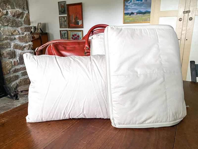 A cream-colored pillow and a zipped cushion on a wooden table, with a red leather bag in the background.