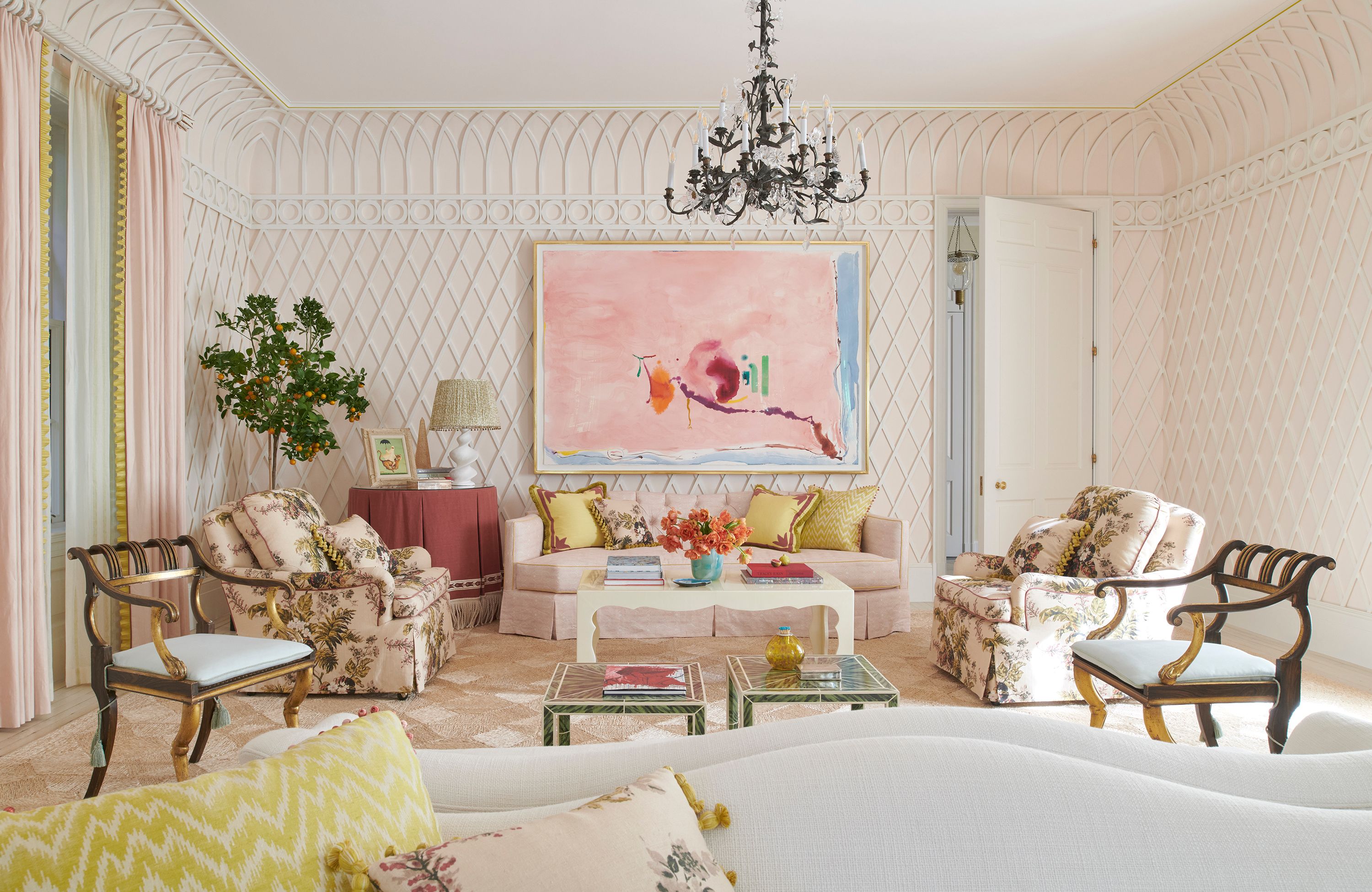 In the living room, giving the appearance of plaster, latticework executed by Fuller Architectural Products curves up to meet the ceiling. A Helen Frankenthaler painting hangs above the Grafton Furniture sofa, which is upholstered in a light pink Studio Four fabric. On other walls, works by Walton Ford and Ed Clark overlook lacquered parchment coffee tables from Etched Design, twin Nickey Kehoe camel-back sofas in Cowtan & Tout cream fabric, and ebonized and parcel gilt Egyptian Revival chairs from the Regency era. Samuel & Sons tassels lend the chairs a playful edge.