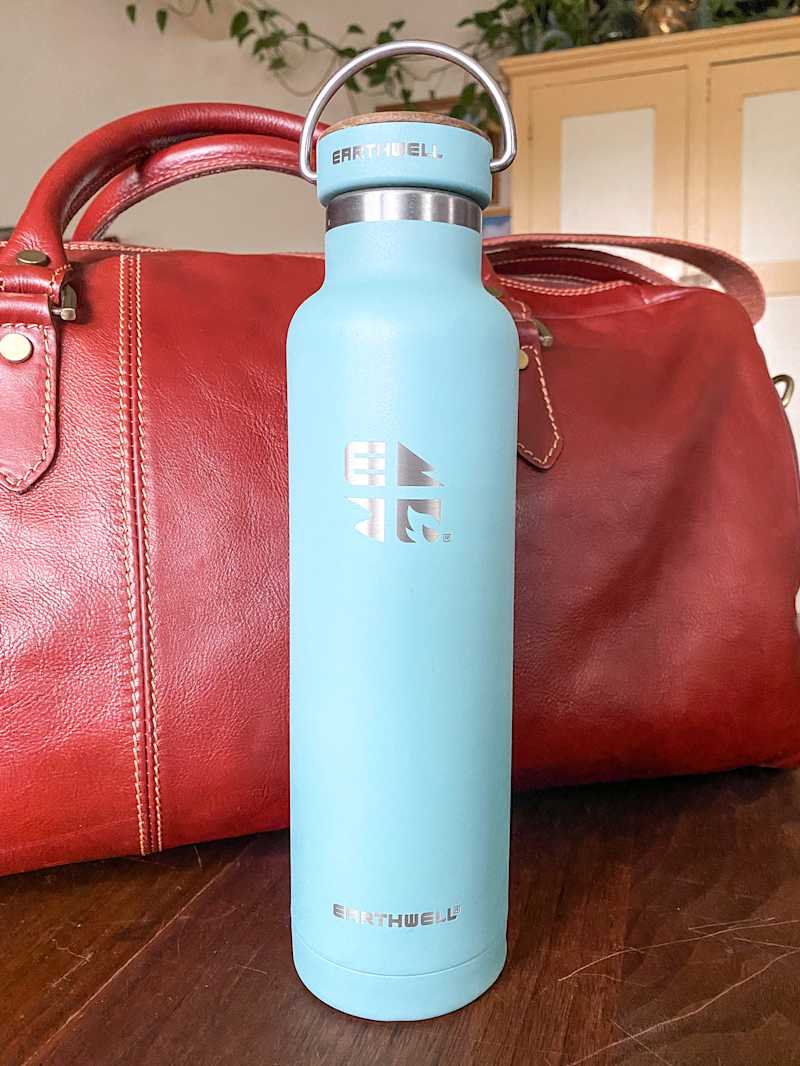 A light blue insulated water bottle with a cork lid next to a red leather bag on a wooden surface.