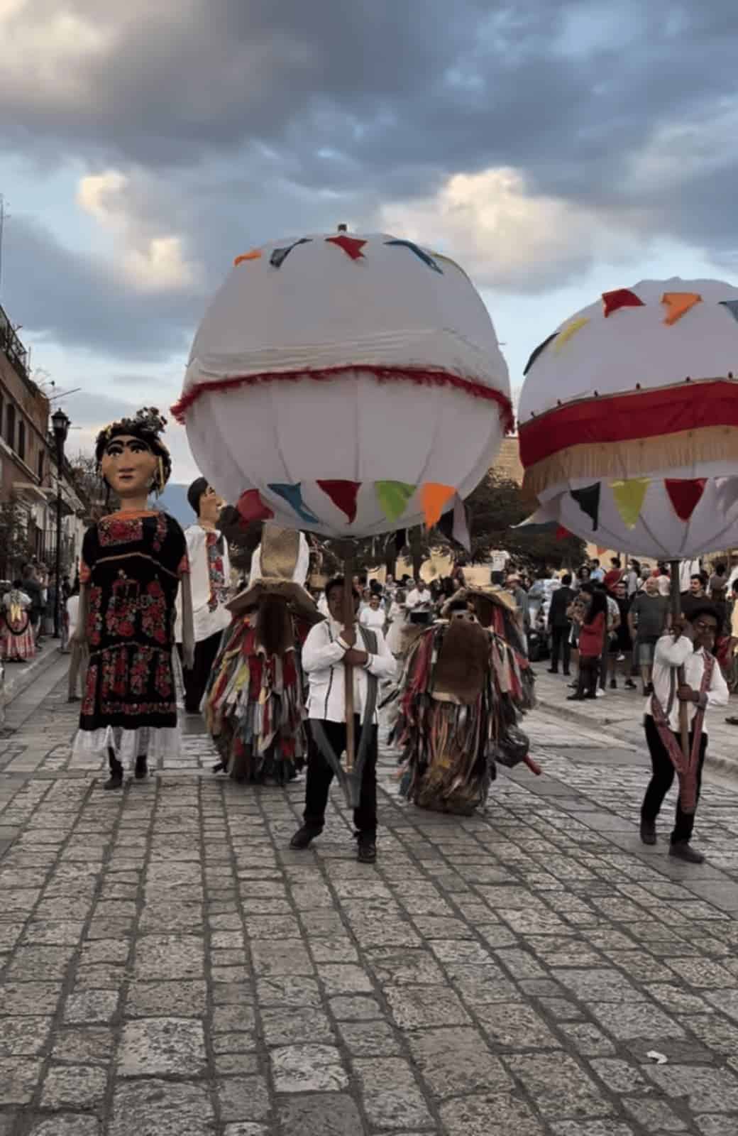 A lively parade moves through a cobblestone street in Oaxaca, featuring towering papier-mâché giant puppets, performers in traditional costumes, and participants carrying oversized white festival lanterns decorated with colorful pennant flags. Crowds line the street beneath dramatic evening clouds, creating a festive atmosphere during a local cultural celebration.