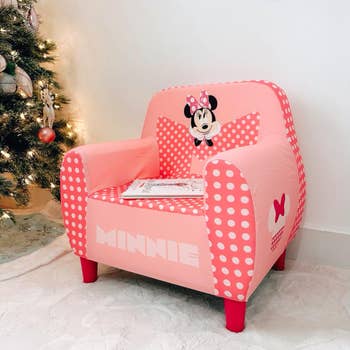 Pink polka-dot children's chair with Minnie Mouse design, placed by a lit Christmas tree, a small book rests on the seat
